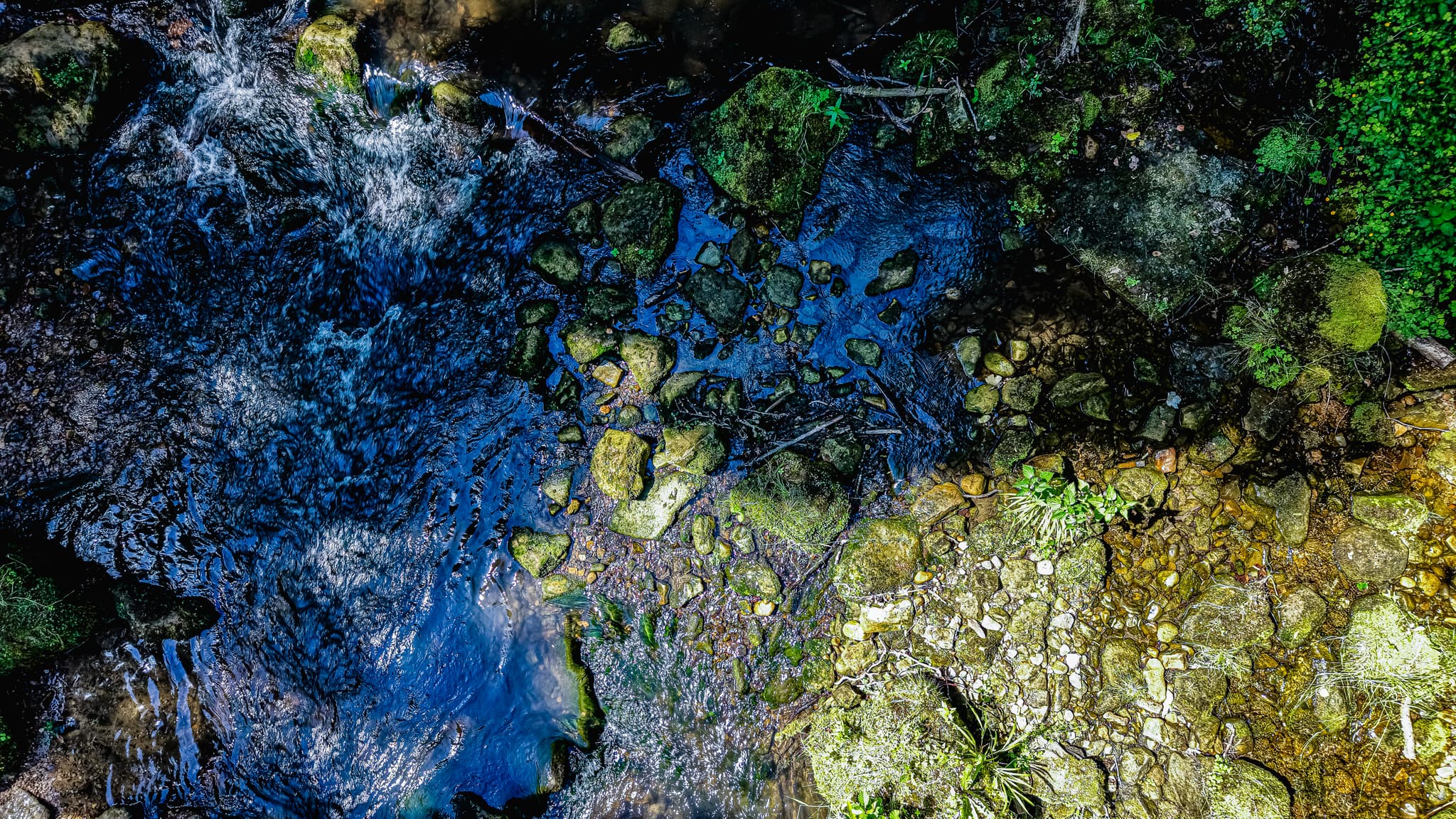 Streambed from directly above — the precise line where shallow amber water gives way to deep blue-black channel. The same river, two depths, two colours. At this altitude the Black Ernz reads as an abstract.