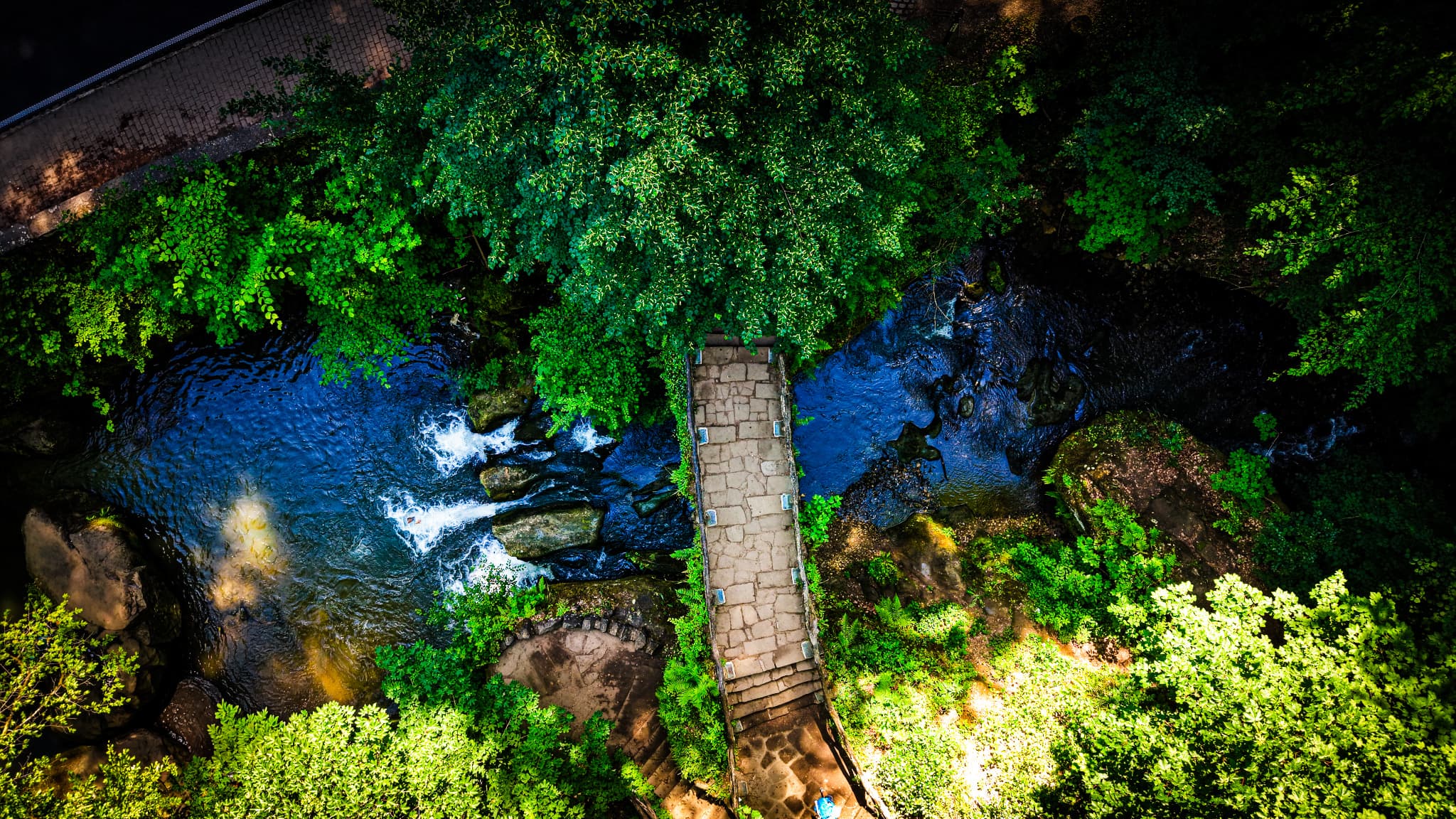 The ancient stone bridge from directly above. From the ground the bridge frames the water. From twenty metres up the relationship inverts — the arch becomes a geometric subject, the blue water its setting. The walkers below cannot have this view.