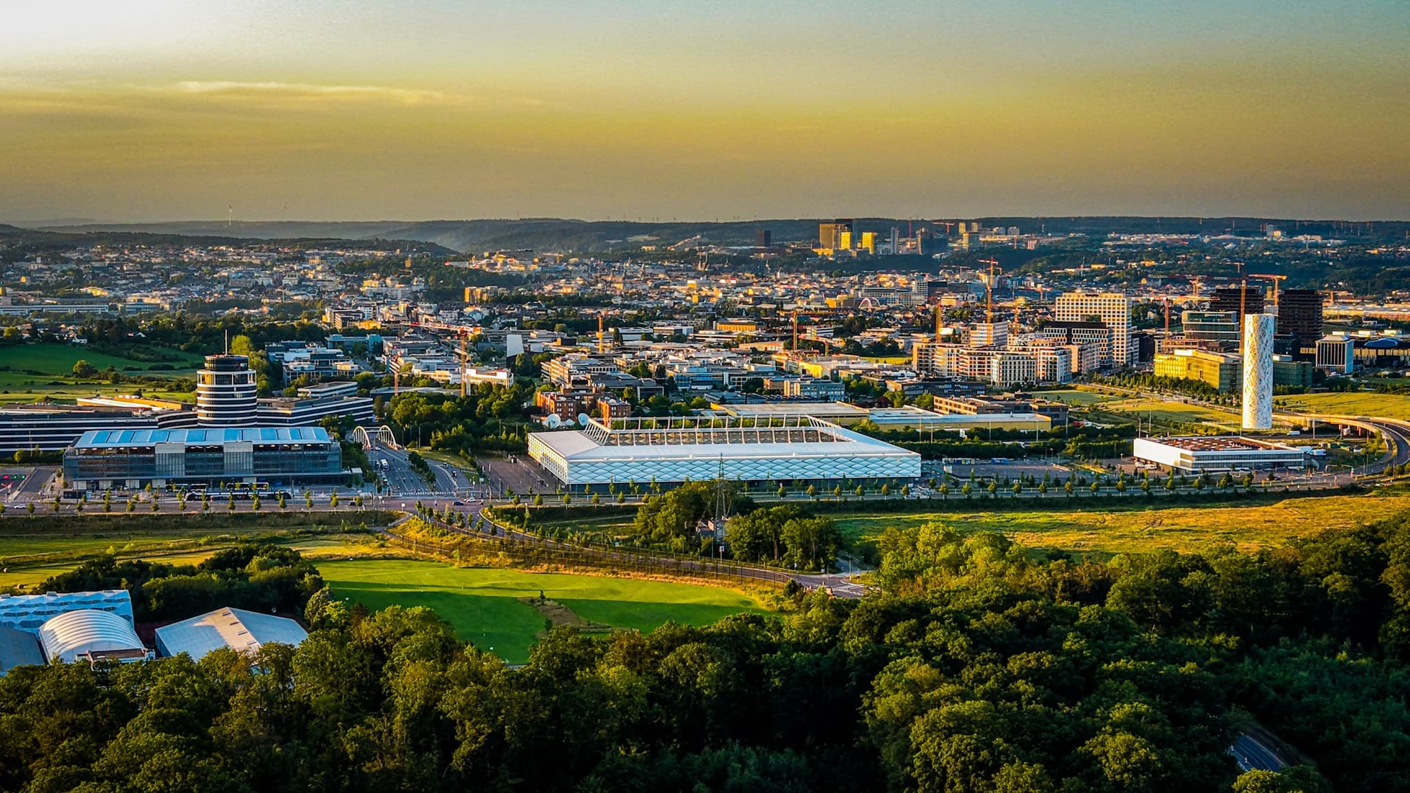 Gasperich in the last light. The Cloche d'Or towers glow at the far edge, the suburb spreads into the frame, and beyond the bottom edge the city simply stops. The same ground that reads as retention lake and field geometry from directly above reads, from here, as a city that doesn't quite know it has ended.