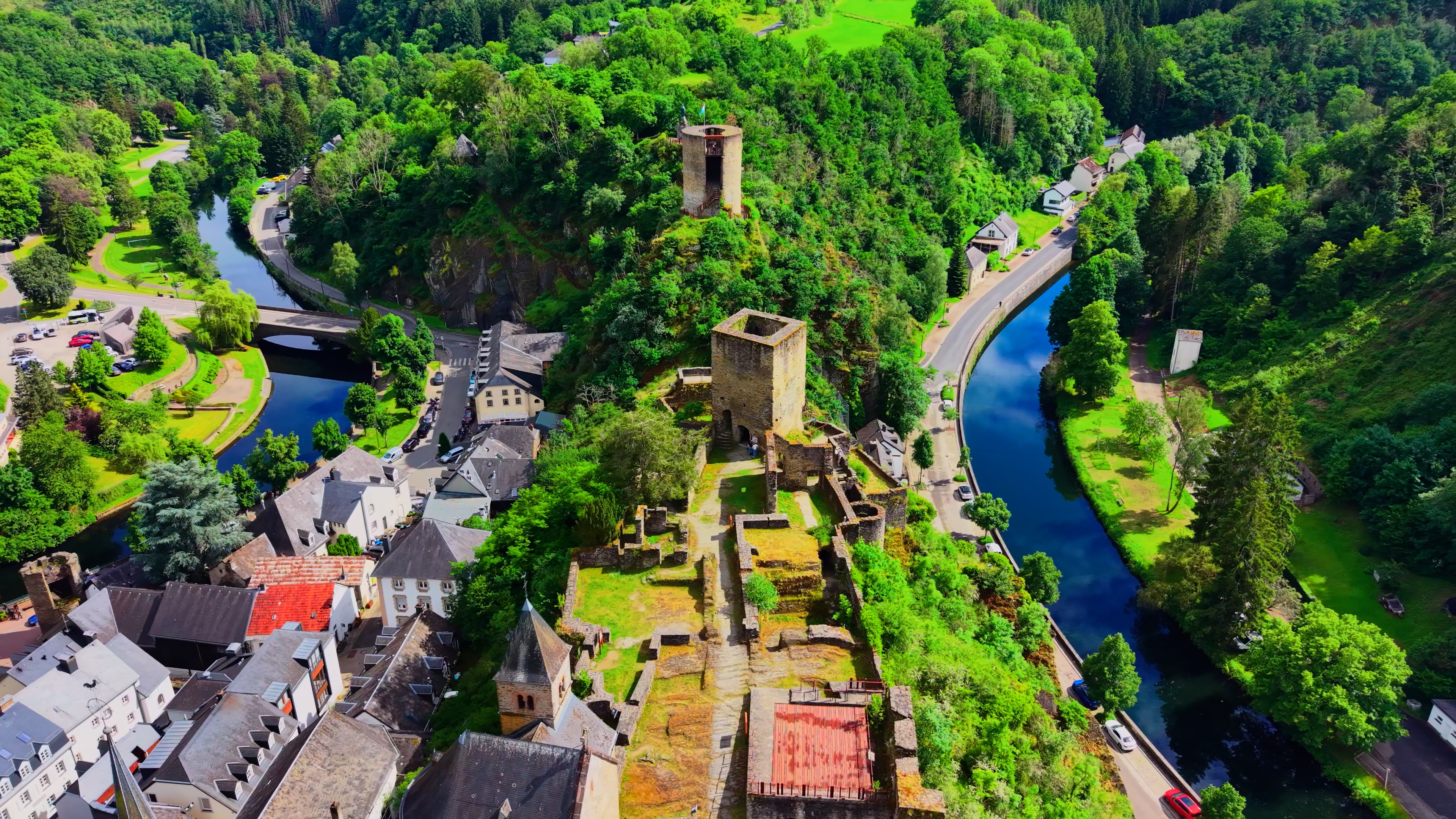 The full castle precinct from altitude — curtain wall, keep, and the Sûre running below on three sides. The village grew up between the bluff and the river, in the only space that was available.