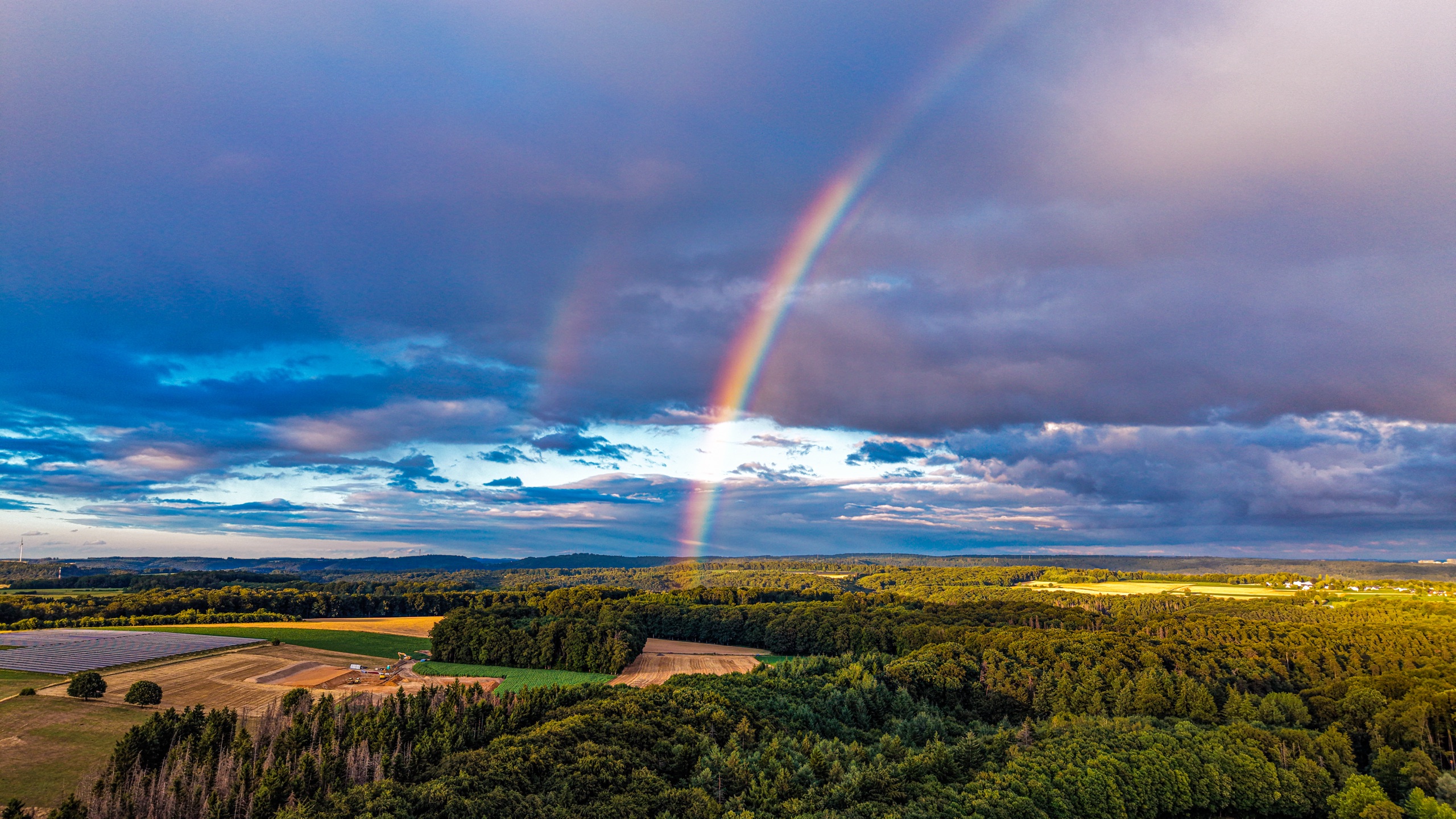 A rainbow touching down in the agricultural fields east of Marienthal — solar panels in the middle ground, wide summer sky
