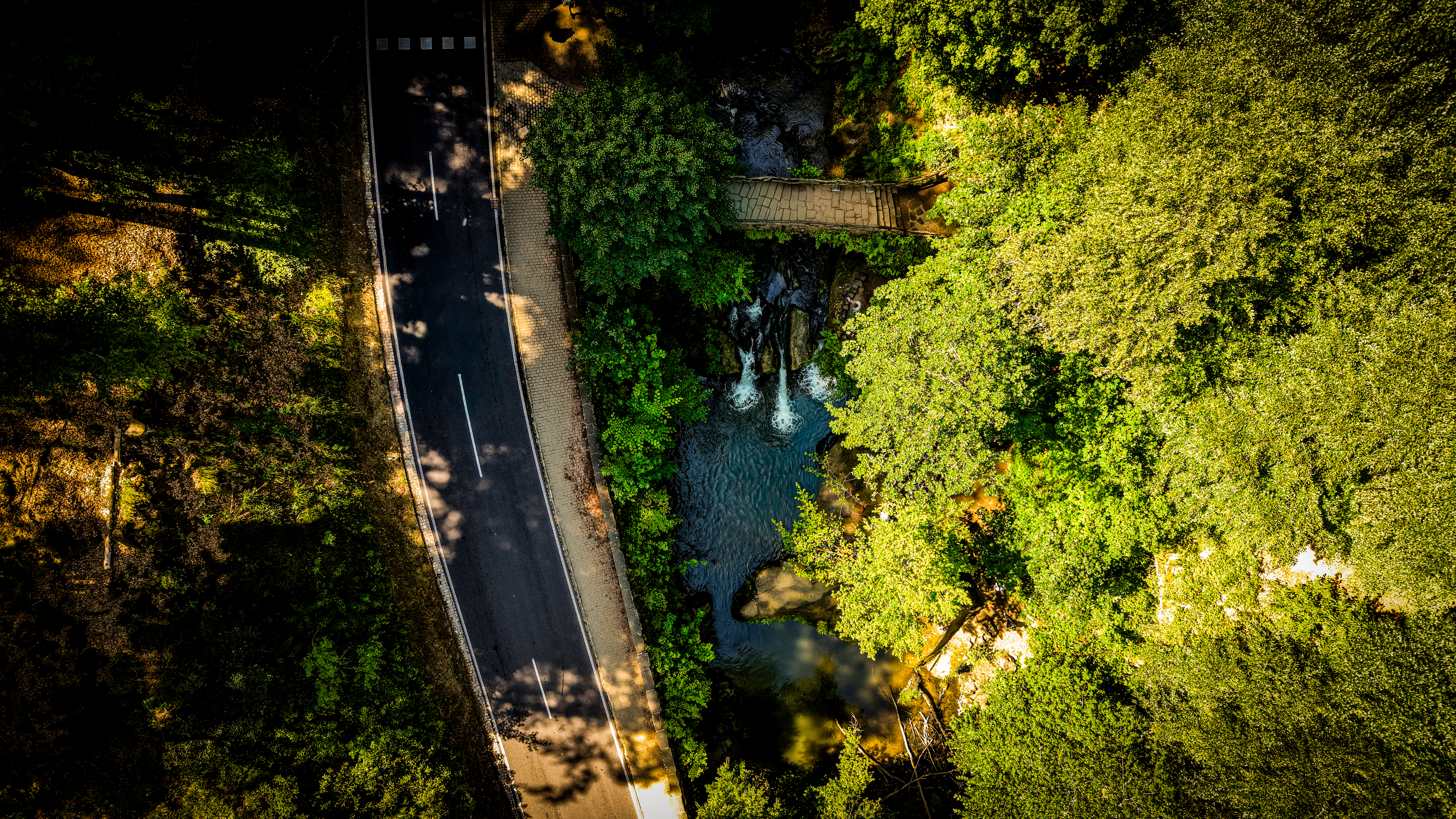 The gorge road from near-nadir — tarmac running straight through the tree canopy, the Black Ernz and its waterfall visible as a break in the green just beside it. The road and the river have been parallel here since the mills were working.