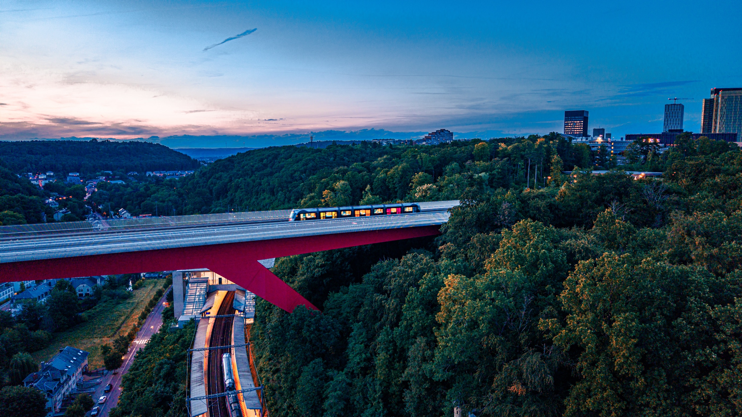 The Pont Rouge at blue hour — a tram crossing the red viaduct, Kirchberg towers lit on the right edge, forest and valley below in deep dusk blue