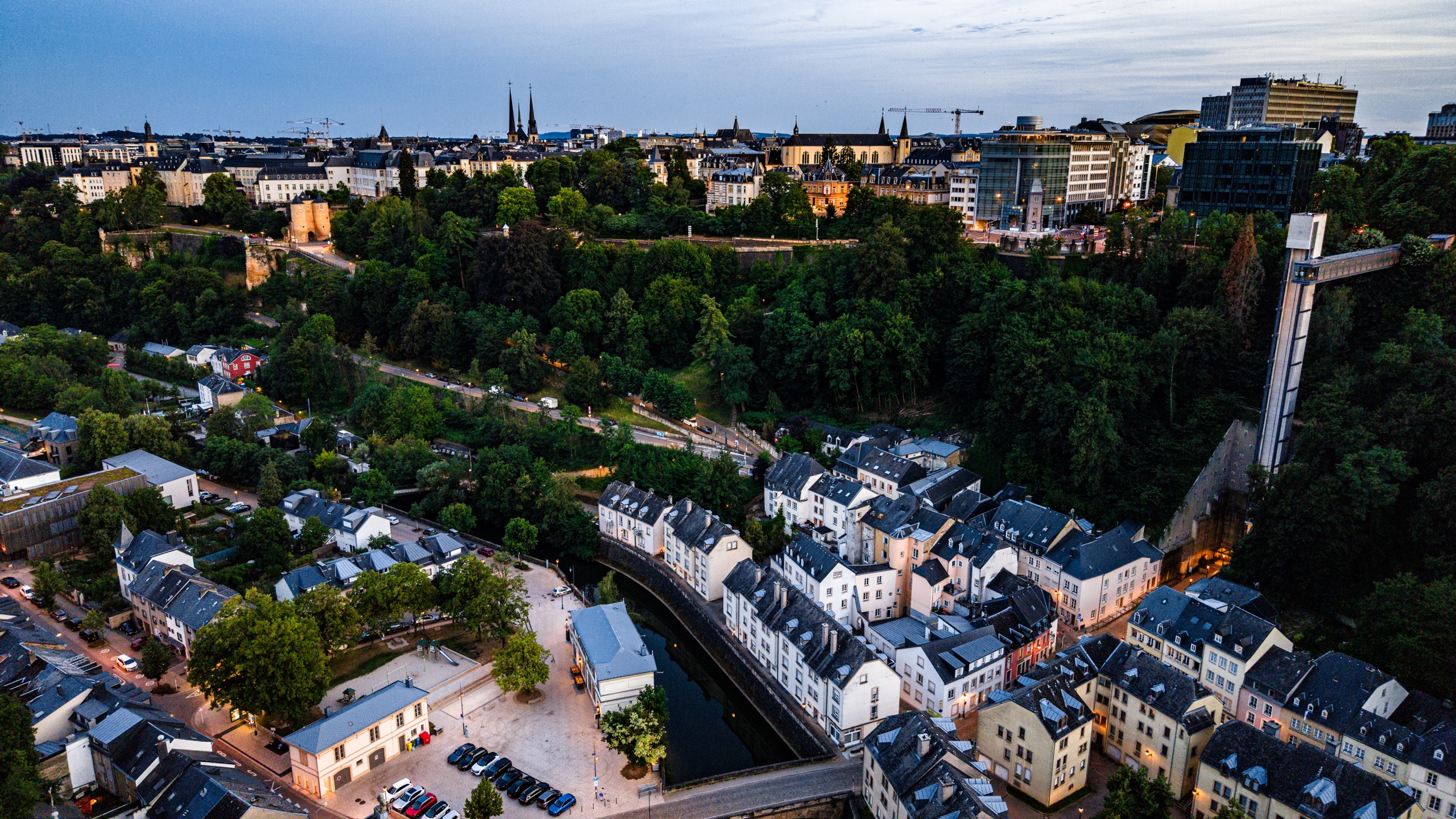 The Grund quarter and Ville Haute from altitude — the Alzette river, Grund's densely packed houses along the water, and the old city on the cliff above with cathedral spires against the fading sky