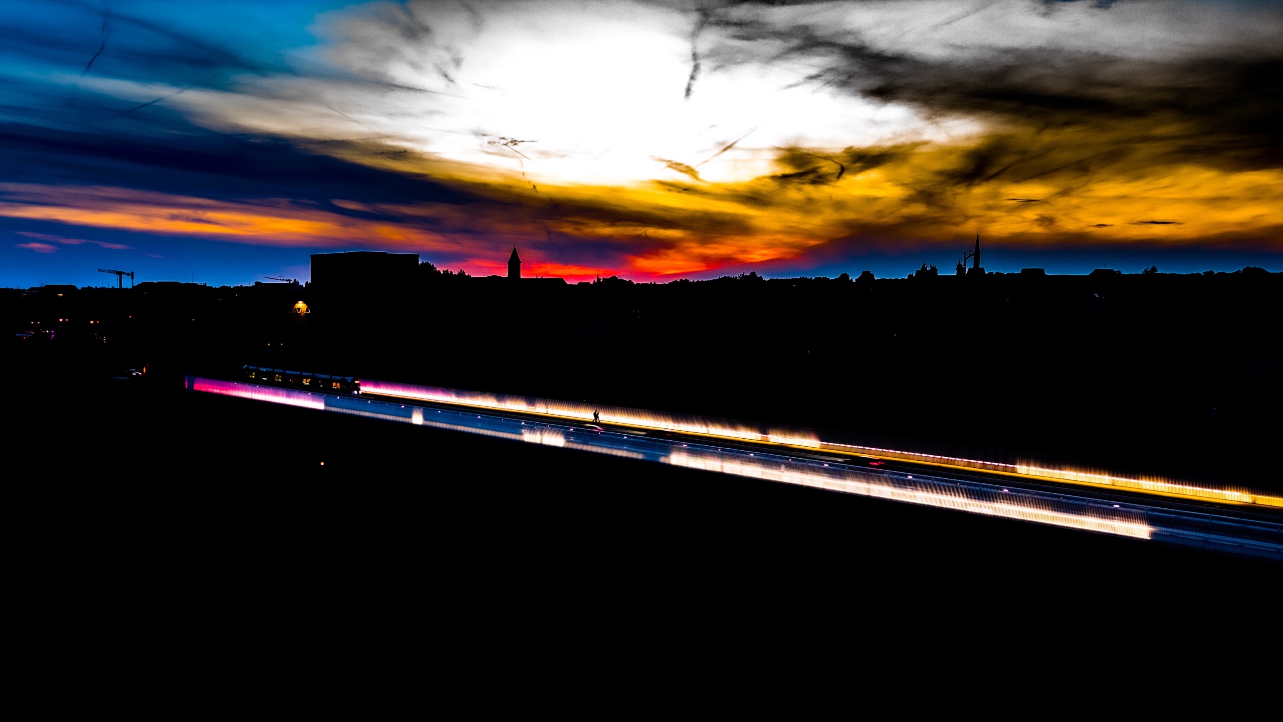 The Pont Rouge bridge deck as a diagonal streak of light — tram blur and LED lighting in purple, gold, and white against a sky of orange fire and dark cloud, church silhouettes at the horizon