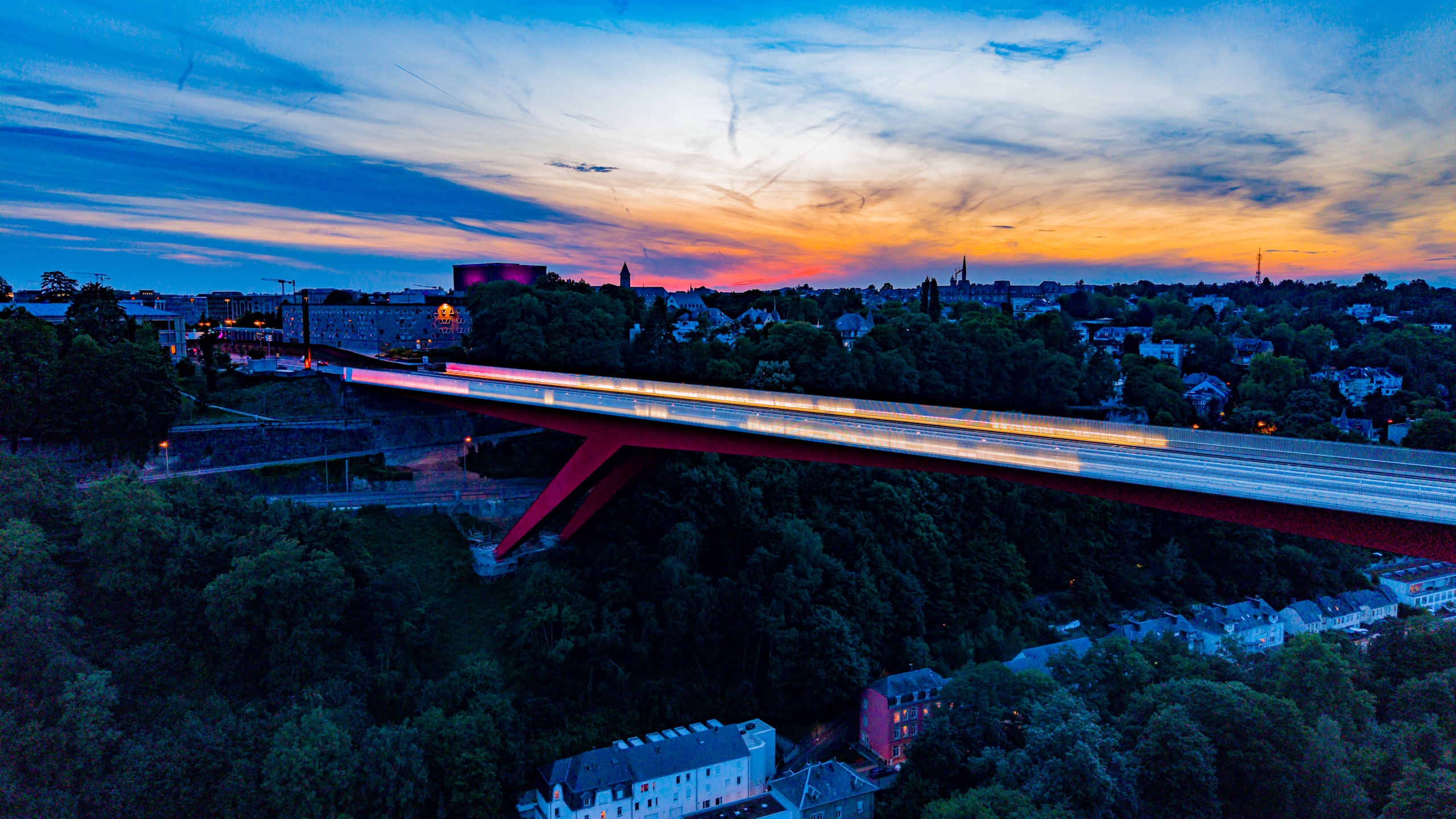 Wide view of the Pont Rouge at dusk — the full red arch against an orange and blue sky, tram blur across the bridge, city on both sides, forest canopy below